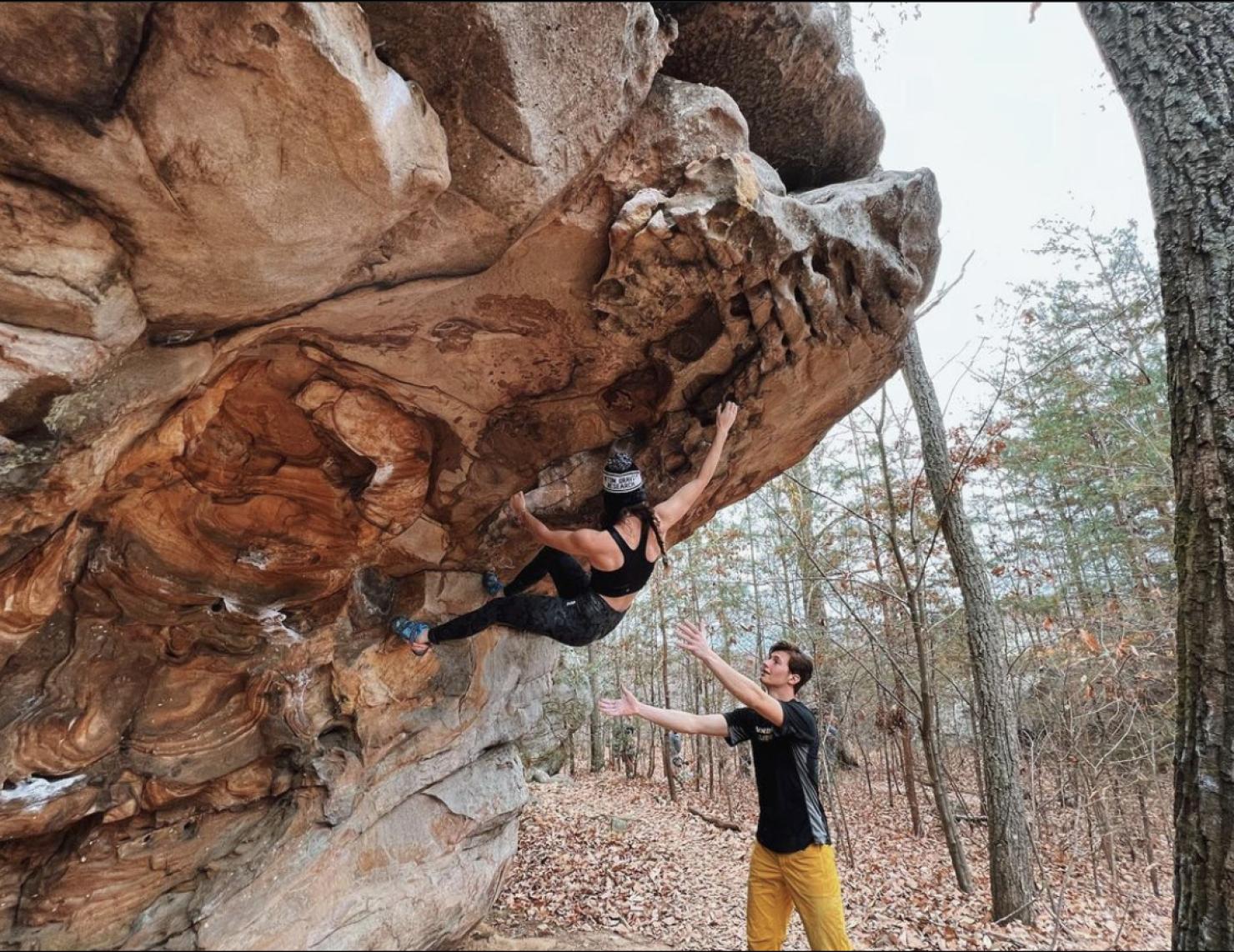 Lauren King climbs a boulder with a spotter on the ground below.