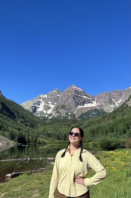 Kylie Yang wearing black sunglasses and a light yellow jacket, hair in two braids, posing in a green meadow with rocky mountains in the background during springtime.