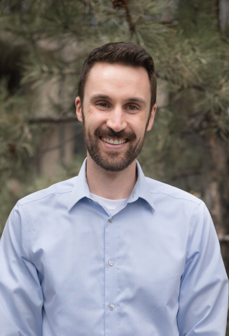 Outdoor headshot photo of a man with brown hair and beard, wearing a baby blue button up. Looks to be taken outside, with pine trees in the background. 