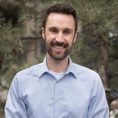 Outdoor headshot photo of a man with brown hair and beard, wearing a baby blue button up. Looks to be taken outside, with pine trees in the background. 