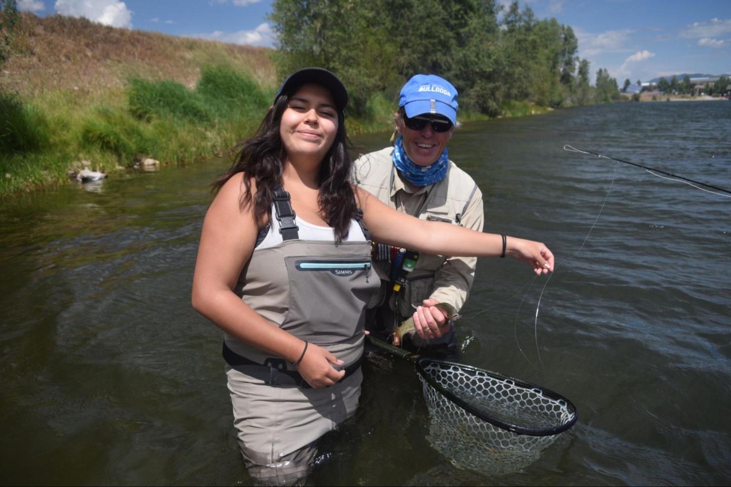 A woman in a river with fly fishing gear, standing in front of an instructor. 