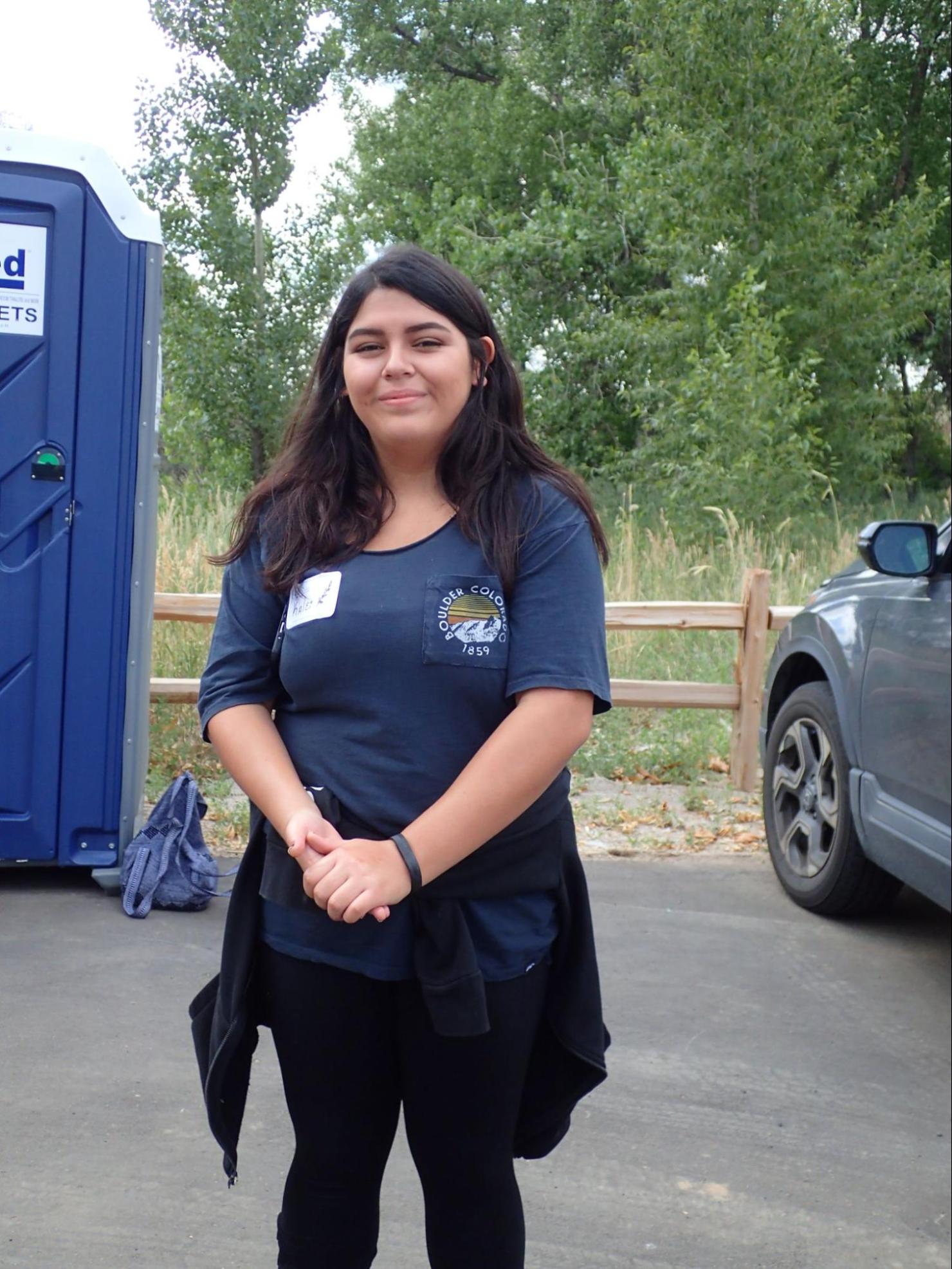 Young woman with dark shoulder length hair smiling for a photo in an outdoor parking lot, presumably near a park with lots of tree coverage.