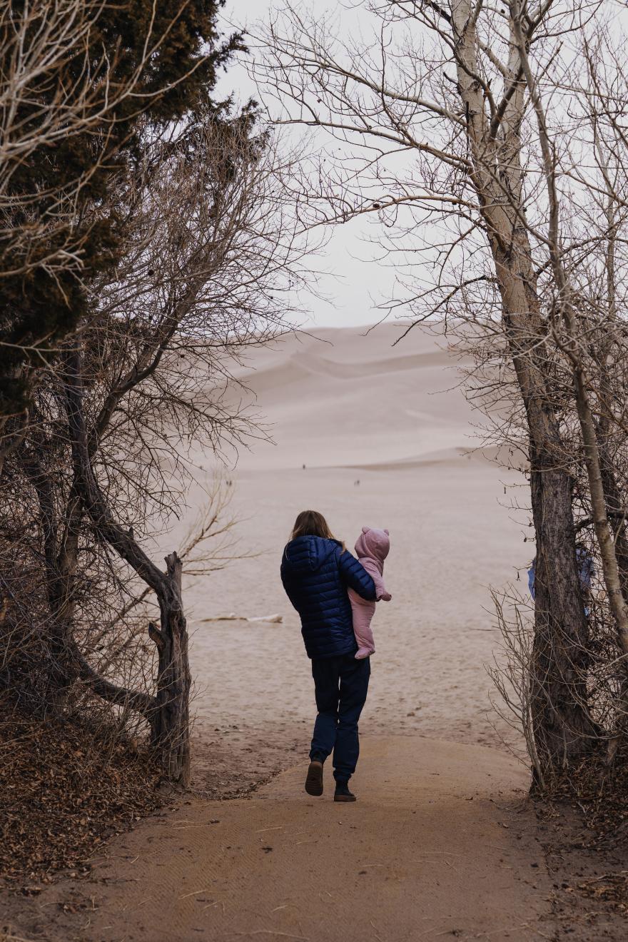 Photo taken from the back of woman holding a child entering sand dunes.