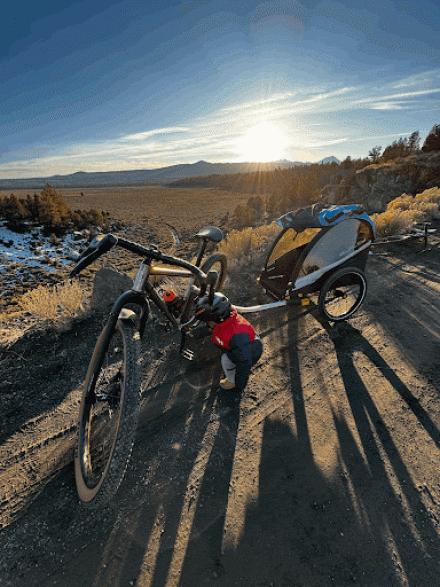 Small child playing with a bike and child trailer on a dirt trail during a sunny day.